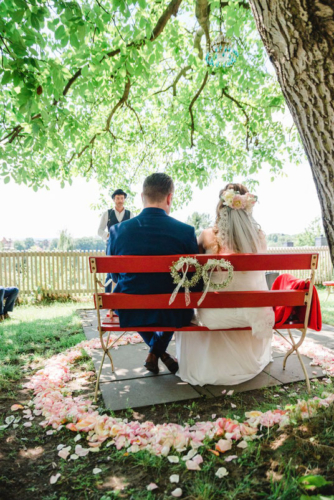 Brautpaar auf einer roten Bank unter Apfelbaum während der freien Trauung